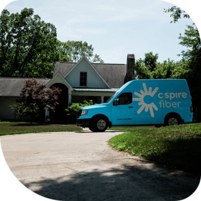 A blue C Spire Fiber van parked on the driveway in front of a suburban house with trees in the background.