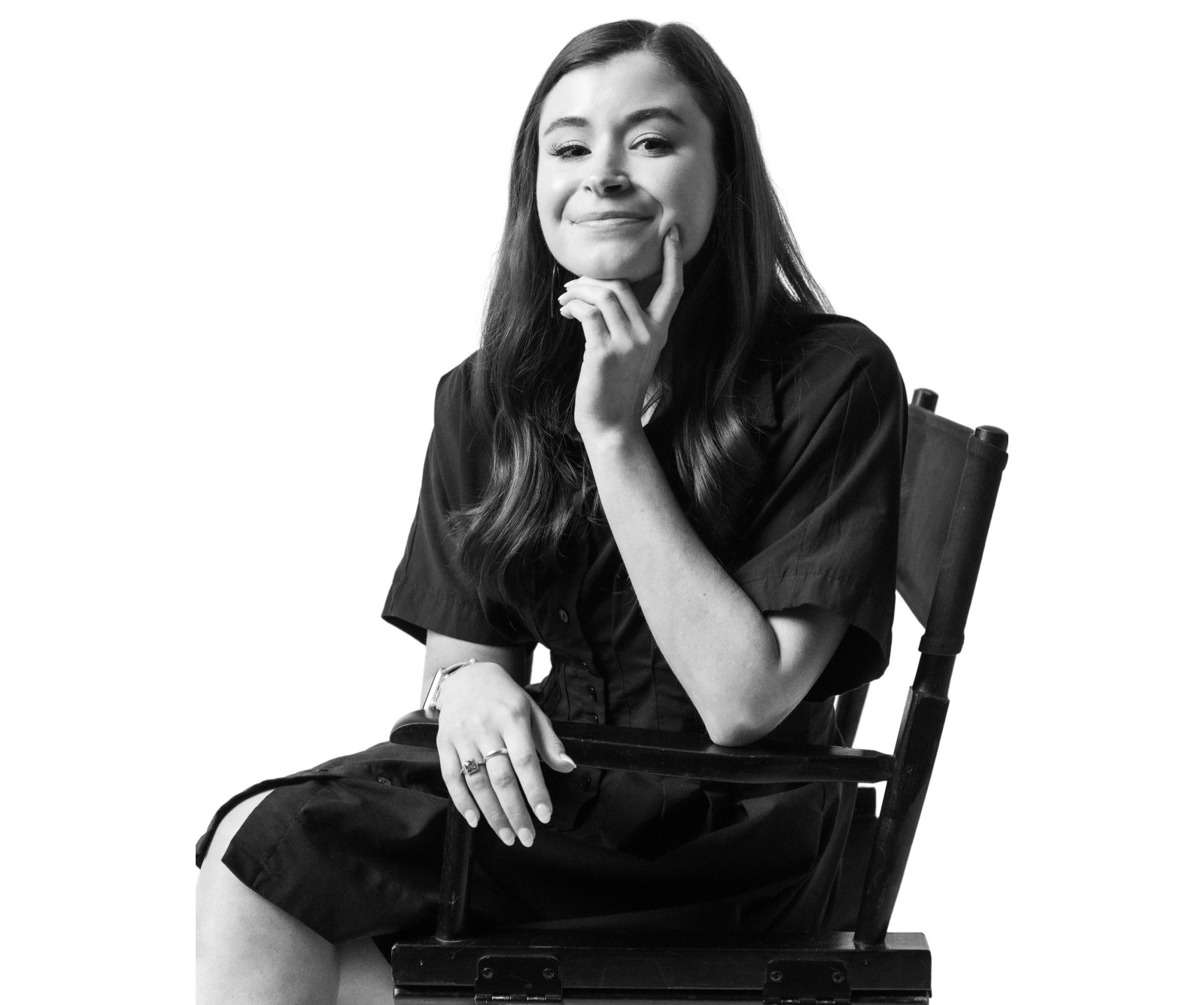 A woman with long hair sits on a chair, smiling slightly and resting her chin on her hand. The image is in black and white.