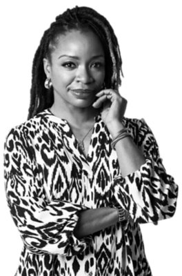 Woman with long, styled hair and hoop earrings wearing a patterned blouse, standing with one arm crossed and the other hand touching her face, looking at the camera.