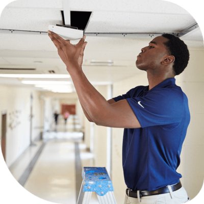 A man in a blue shirt stands on a stepladder, installing or inspecting a device in a ceiling tile in a hallway.
