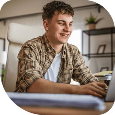 Young man seated at a desk, smiling and typing on a laptop in a home office setting with shelves and plants in the background.