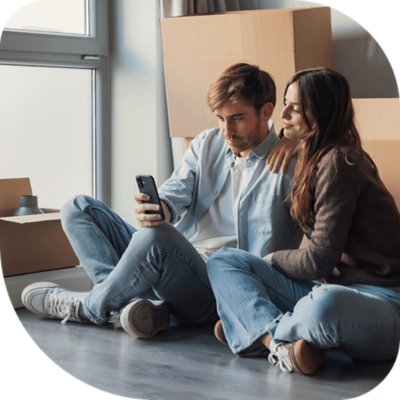 A couple sitting on the floor surrounded by moving boxes, looking at a phone.