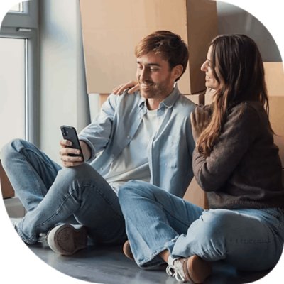 A man and woman sit on the floor surrounded by moving boxes, looking at a phone together near a window as they settle in after moving to a new home.