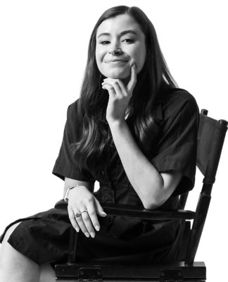 A woman with long hair sits on a chair, smiling slightly and resting her chin on her hand. The image is in black and white.